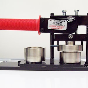 May include: A black button maker with a red handle. The machine has two metal cylinders and a black base. The text on the machine reads "BUTTON MAKER MODEL 125 Patent Nos. issued 5,374,044 & 5,471,617 Manufactured in the USA by TECRE POND DU LAC, WI."