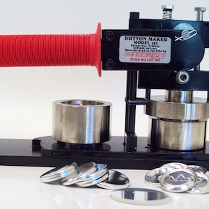 May include: A black button maker with a red handle. The machine is used to create round buttons. There are several silver buttons on the table in front of the machine.