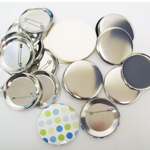 May include: A collection of round, silver-coloured metal button blanks, some with safety pins attached. There is also a white round blank and a button with a blue and green polka dot design.