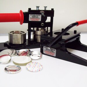 Op de afbeelding: Twee zwarte knoopmachines met rode handvatten. De machine aan de linkerkant is gelabeld "Button Maker Model 17A" en de machine aan de rechterkant is gelabeld "Circle Punch Model 208A". Er liggen verschillende ronde metalen en plastic knoopblanks rond de machines.