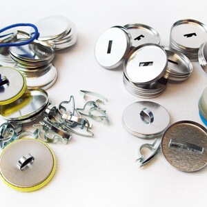 Op de afbeelding: Assortiment van zilveren metalen knoopsmaakbenodigdheden, waaronder achterkanten, voorkanten en een blauw elastiek. Er zijn ook verschillende zilveren metalen pin-achterbevestigingen.