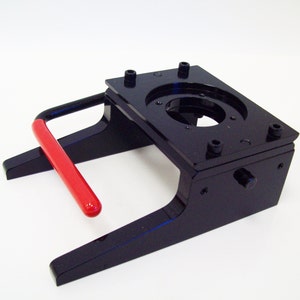 May include: Black metal button maker with a red handle. The button maker has a round opening for inserting a button.