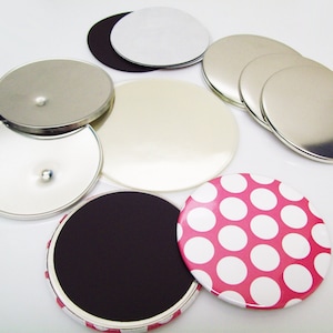 May include: A collection of round, silver-colored metal button blanks and mirrors. Some have a shiny, reflective surface, while others are matte. There are also a few with a white or black backing, and one with a pink and white polka dot design.