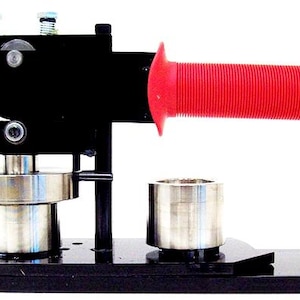 May include: A black metal button maker with a red handle. The machine has a metal base and a metal cylinder for making buttons.