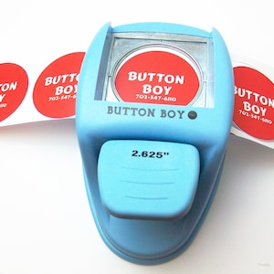 May include: A blue button maker with a 2.625 inch diameter circle die. The machine is labeled "Button Boy" and has a red circle sticker with the text "Button Boy" and a phone number.