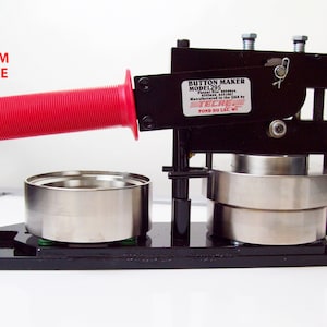 Op de afbeelding: Een zwarte knopenmaker met een rode handgreep. De machine heeft twee metalen cilinders en een label met de tekst "BUTTON MAKER MODEL 295 Patent No. 6078944 Manufactured in the USA by TECRE FOND DU LAC WI".