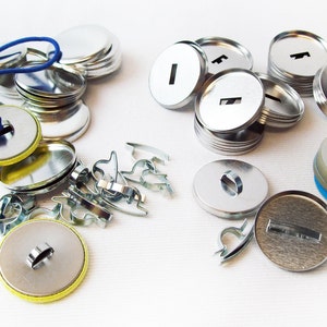 Op de afbeelding: Assortiment van zilveren metalen knoopsmaakbenodigdheden, waaronder achterkanten, voorkanten en een blauw elastiek. Er zijn ook verschillende zilveren metalen pin-achterbevestigingen.