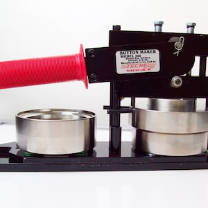 May include: Black button maker with a red handle. The machine has two metal cylinders and a label that reads "Button Maker Model 300 Patent No. 5,930,987 Manufactured in the USA by TECHE  FOND DU LAC, WI."