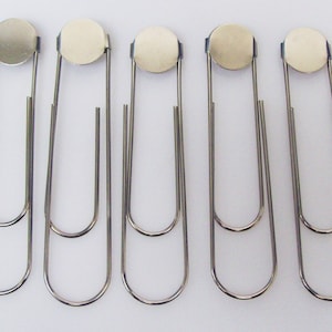May include: Five silver-coloured metal paper clips with a circular top and a double-loop design. The clips are arranged in a row against a white background. Each clip has a shiny, reflective surface.