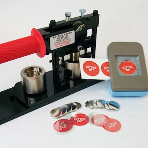 Op de afbeelding: Een zwarte knopenmaker met een rode handgreep, een grijze knopenpons en een vel ronde stickers met de tekst "Button Boy". Er zijn ook verschillende zilveren knoopruggen en rode knoopvoorkanten.