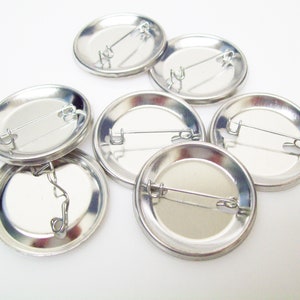 Puede incluir: Una colección de botones de solapa en blanco, de color plateado. Cada botón tiene un diseño circular con un cierre tipo imperdible en la parte posterior. Los botones están dispuestos sobre una superficie blanca.