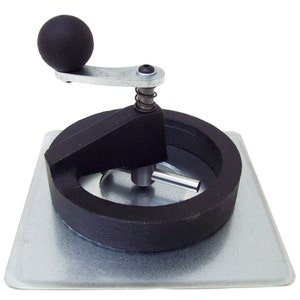May include: A black metal circle cutter with a handle and a spring. The cutter is on a silver metal base.