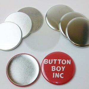 Op de afbeelding: Een set zilveren metalen knoopsluitingen en een rode knop met de tekst "Button Boy Inc."