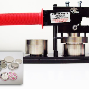 May include: A black button maker with a red handle. The machine is made of metal and has a silver base. The machine is used to make buttons with a diameter of 1.25 inches. The machine is made by ECKERT. The machine is also shown with a variety of buttons, including blank buttons and buttons with a floral design.