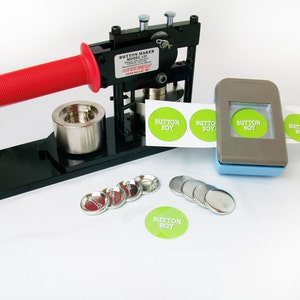 May include: A black button maker with a red handle, a gray button punch, and a sheet of green and white button stickers that say "Button Boy". There are also several silver button backs and fronts.