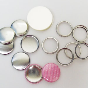 May include: A set of silver button making supplies, including backings, fronts, and a white circle for a button design. There is also a pink button with white polka dots.