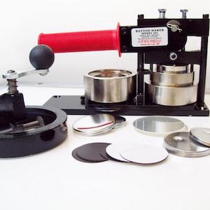 May include: A black button maker machine with a red handle and a silver base. The machine is used to create custom buttons. The machine is surrounded by various button parts, including metal bases, plastic covers, and paper inserts.