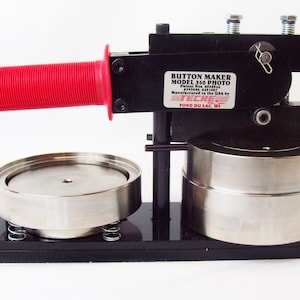 May include: A black button maker with a red handle. The machine is labeled "Button Maker Model 350 Photo" and "Tecre".