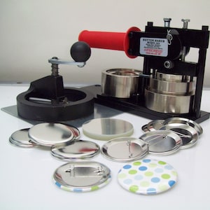 May include: A black button maker machine with a red handle and a silver base. The machine is surrounded by a variety of blank and decorated buttons.