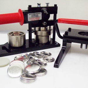 May include: A black button maker with a red handle, a silver base, and a separate black base with a silver cylinder. The machine is surrounded by a pile of silver button parts.