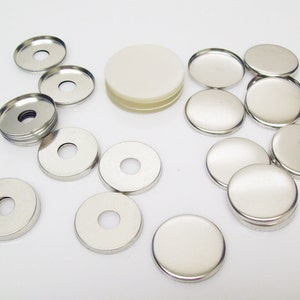 May include: Assortment of silver metal button blanks in various sizes. The blanks are round and have a hole in the center. Some of the blanks are stacked on top of each other.