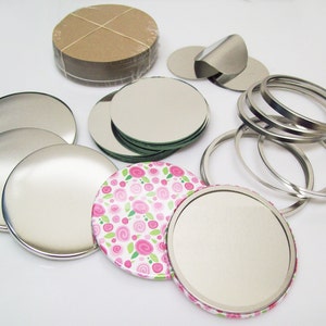 May include: Assortment of round metal button blanks and mirror blanks in various sizes. The blanks are silver in color and some have a pink floral design. There are also several round metal rings.