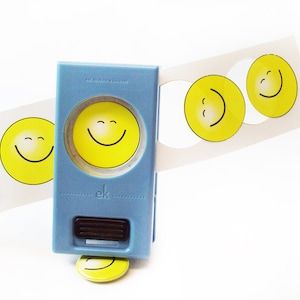 May include: A blue and white paper punch with a smiley face design. The punch is used to create small, round smiley face stickers. The punch is sitting on a white surface with a sheet of paper with several smiley face stickers.