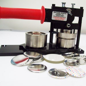 May include: A black button maker machine with a red handle. The machine has two metal cylinders and a base. There are several round metal and plastic buttons in various sizes and colors scattered around the machine.