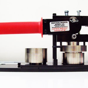 May include: A black button maker with a red handle. The machine is labeled "Button Maker Model 150 Patent No. 6,030,864  6,335,040  Manufactured in the USA by ECRE  FOND DU LAC, WI."