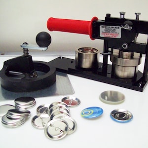 May include: A black button maker with a red handle, a silver die, and a silver base. The machine is on a white surface with a variety of silver button blanks, a clear plastic button blank, and a blue and white button with a design.