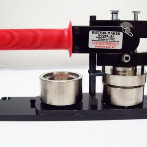 Op de afbeelding: Een zwarte knopenmaker met een rode handgreep. De machine is gelabeld "BUTTON MAKER MODEL 175 Patent No. 6039844 Manufactured in the USA by TECRE FOND DU LAC, WI."