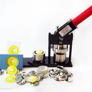 May include: A black button maker with a red handle, a clear plastic mold, and a pile of silver button backs. There are also several yellow smiley face buttons and a blue plastic button maker.