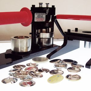 May include: A black button maker machine with a red handle and a silver base. The machine is used to create round buttons with a diameter of 1 inch. The machine is shown with a variety of button parts, including backs, fronts, and pins.