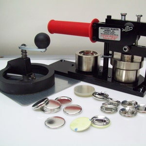 May include: A black button maker with a red handle, silver components, and a black base. The machine is labeled "Button Maker Model 185". Several silver button components and finished buttons are scattered around the machine.