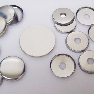May include: A set of silver metal button blanks for making custom buttons. The set includes both round and circular blanks with a hole in the center. There is also a white blank button.