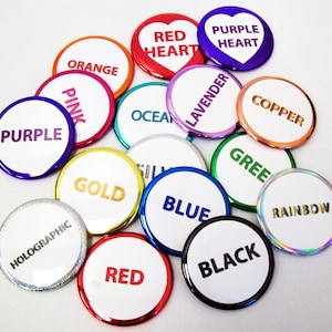 May include: Assortment of colorful round buttons with various words and phrases, including "Red Heart", "Purple Heart", "Orange", "Pink", "Ocean", "Lavender", "Copper", "Purple", "Gold", "Blue", "Green", "Rainbow", "Holographic", "Red", and "Black".