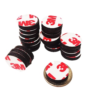 May include: A collection of black, circular adhesive pads with white and red "3M" branding. The pads are stacked in several piles, with one pad resting on a coin for size comparison. The adhesive side is visible.