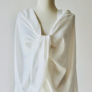 Puede incluir: Una bufanda blanca y ligera hecha de una tela suave y fluida. La bufanda está drapeada sobre un maniquí y anudada en un nudo flojo en el cuello.