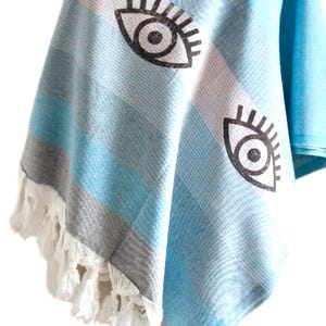 Puede incluir: Una toalla de playa a rayas azul claro y gris con flecos blancos. La toalla tiene un diseño de ojo negro y blanco impreso.
