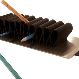 Puede incluir: Un porta l&aacute;pices de espuma negra con una base de metal y una tira de espuma negra en el lateral. El porta l&aacute;pices tiene m&uacute;ltiples ranuras para l&aacute;pices y est&aacute; dise&ntilde;ado para mantener los l&aacute;pices en posici&oacute;n vertical. Hay tres l&aacute;pices en el porta l&aacute;pices, uno marr&oacute;n y dos azules.