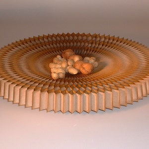 May include: A circular, wooden bowl with a unique, geometric design. The bowl is made of thin, overlapping pieces of wood that create a radial pattern. The bowl is filled with a small pile of peanuts.