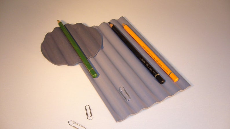 Puede incluir: Un porta l&aacute;pices gris corrugado con tres l&aacute;pices: un l&aacute;piz verde, un l&aacute;piz negro y un l&aacute;piz amarillo. Tambi&eacute;n hay tres clips de papel plateados sobre una superficie blanca.