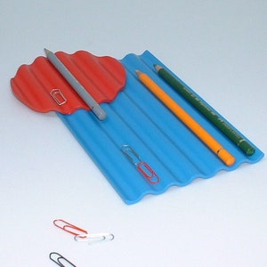 Puede incluir: Un organizador de escritorio azul y rojo con bordes ondulados. El organizador tiene tres ranuras para l&aacute;pices y una peque&ntilde;a bandeja roja para clips. Hay tres clips en el escritorio.