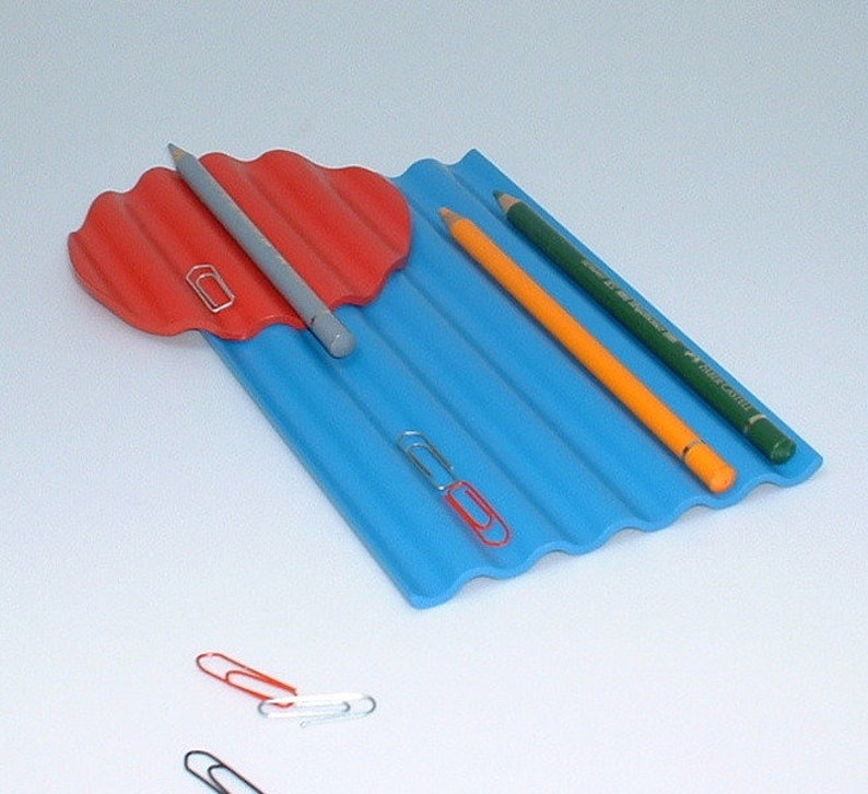 Puede incluir: Un organizador de escritorio azul y rojo con bordes ondulados. El organizador tiene tres ranuras para l&aacute;pices y una peque&ntilde;a bandeja roja para clips. Hay tres clips en el escritorio.