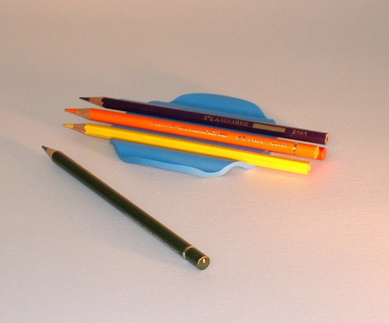 Puede incluir: Cuatro l&aacute;pices de colores en un porta l&aacute;pices azul. Los l&aacute;pices son morado, naranja, amarillo y verde. El porta l&aacute;pices tiene forma de ola.