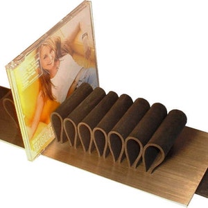 Puede incluir: Un soporte de CD de cuero marr&oacute;n con una caja de CD que presenta a una mujer en la portada. El soporte de CD est&aacute; hecho de m&uacute;ltiples bucles de cuero y est&aacute; sentado sobre una superficie de madera.