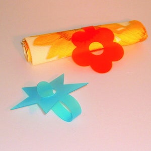 Op de afbeelding: Twee servetringen, een in de vorm van een bloem en de andere in de vorm van een ster. De bloemring is rood en de sterring is blauw.