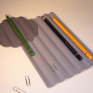 Op de afbeelding: Een grijze, gegolfde potloodhouder met drie potloden: een groen potlood, een zwart potlood en een geel potlood. Er zijn ook drie zilveren paperclips op een wit oppervlak.