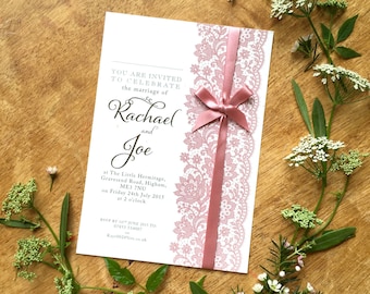 Puede incluir: Una invitación de boda blanca con ribete de encaje rosa y una cinta de satén rosa atada en un lazo. La invitación dice "Están invitados a celebrar el matrimonio de Rachael y Joe" e incluye la fecha, la hora y el lugar de la boda.