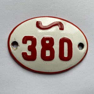 Peut inclure: Plaque ovale en métal vintage, fond blanc et bordure rouge. Le chiffre "380" est affiché en rouge, avec un symbole "S" rouge stylisé. Deux petits trous sont visibles pour le montage.